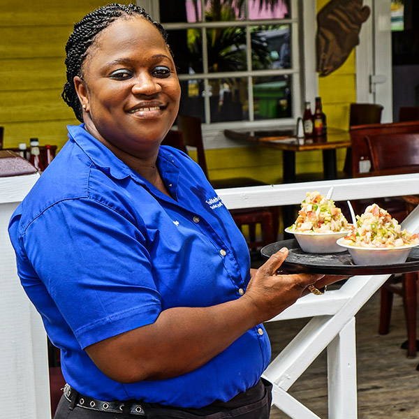 A smiling server carrying two fresh conch salads to the porch tables