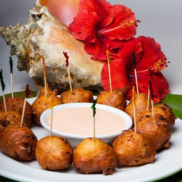 Conch fritters on picks around a bowl of pink fritter sauce, with a conch shell
